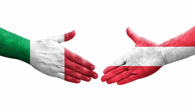 2 sich reihende Hände mit der österreichischen bzw. italienischen Flagge darauf.